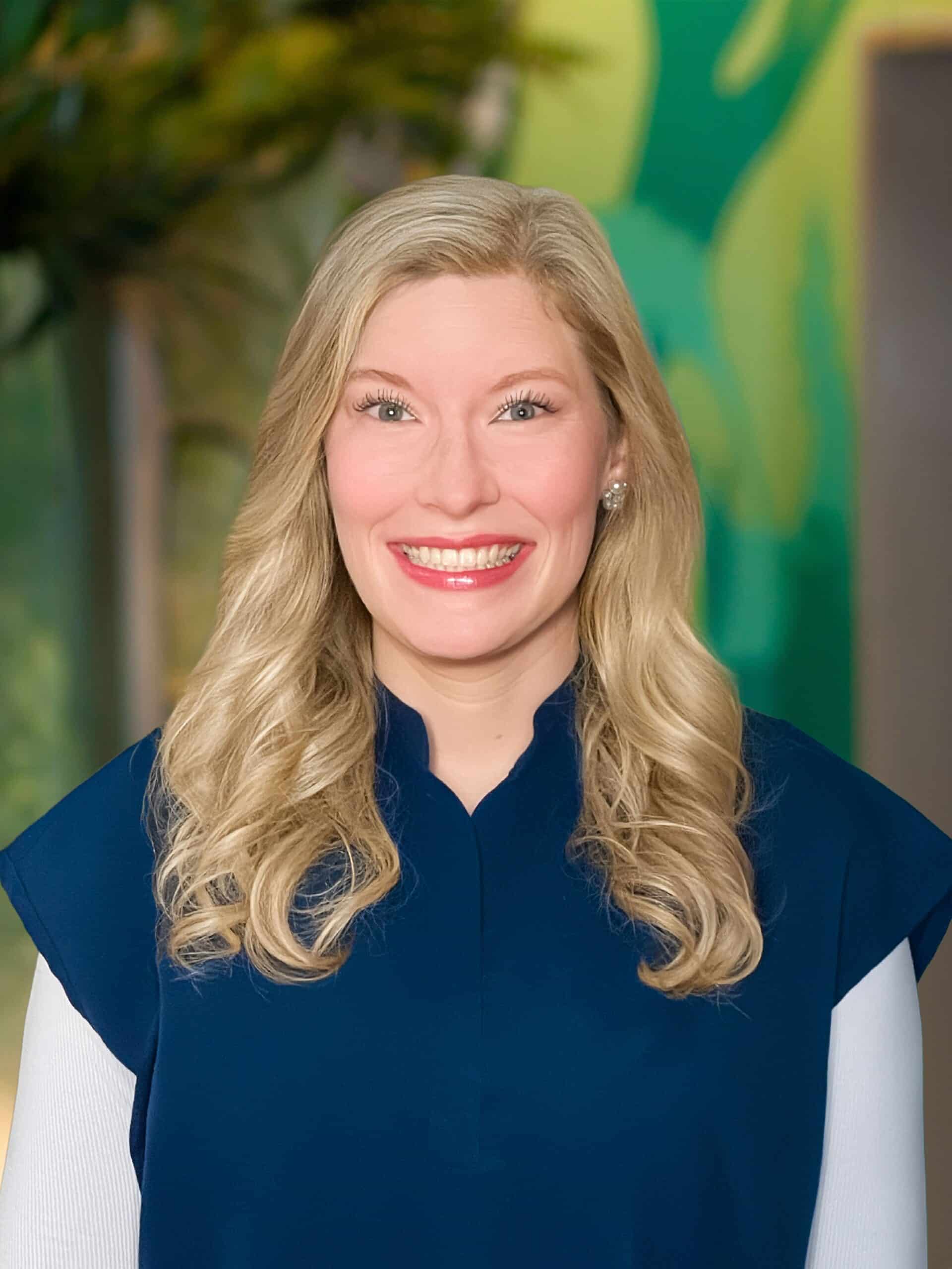 Smiling woman with long blonde hair wearing a dark blue top and white sleeves, neutral colorful background behind her.
