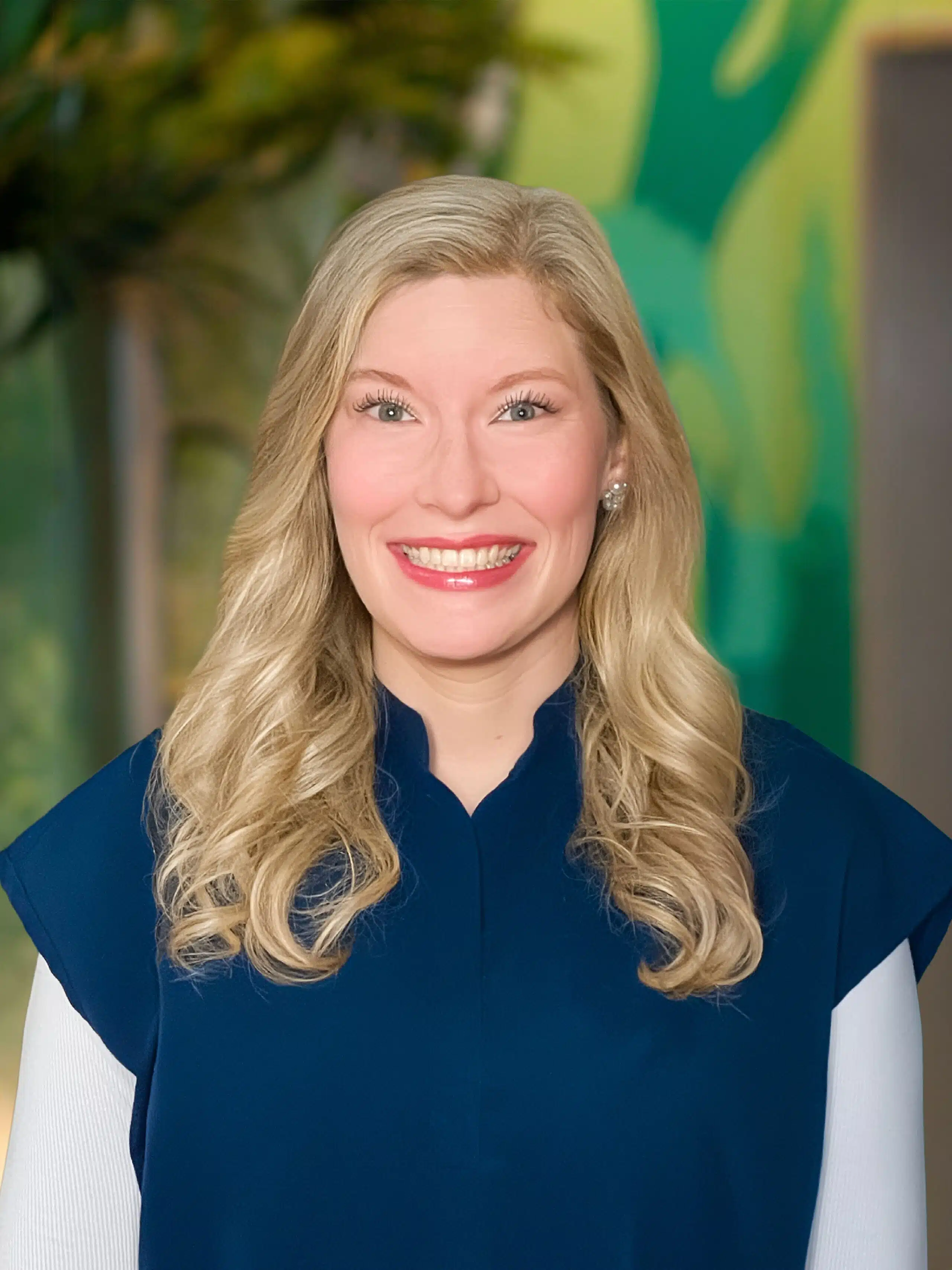 Smiling woman with long blonde hair wearing a dark blue top and white sleeves, neutral colorful background behind her.