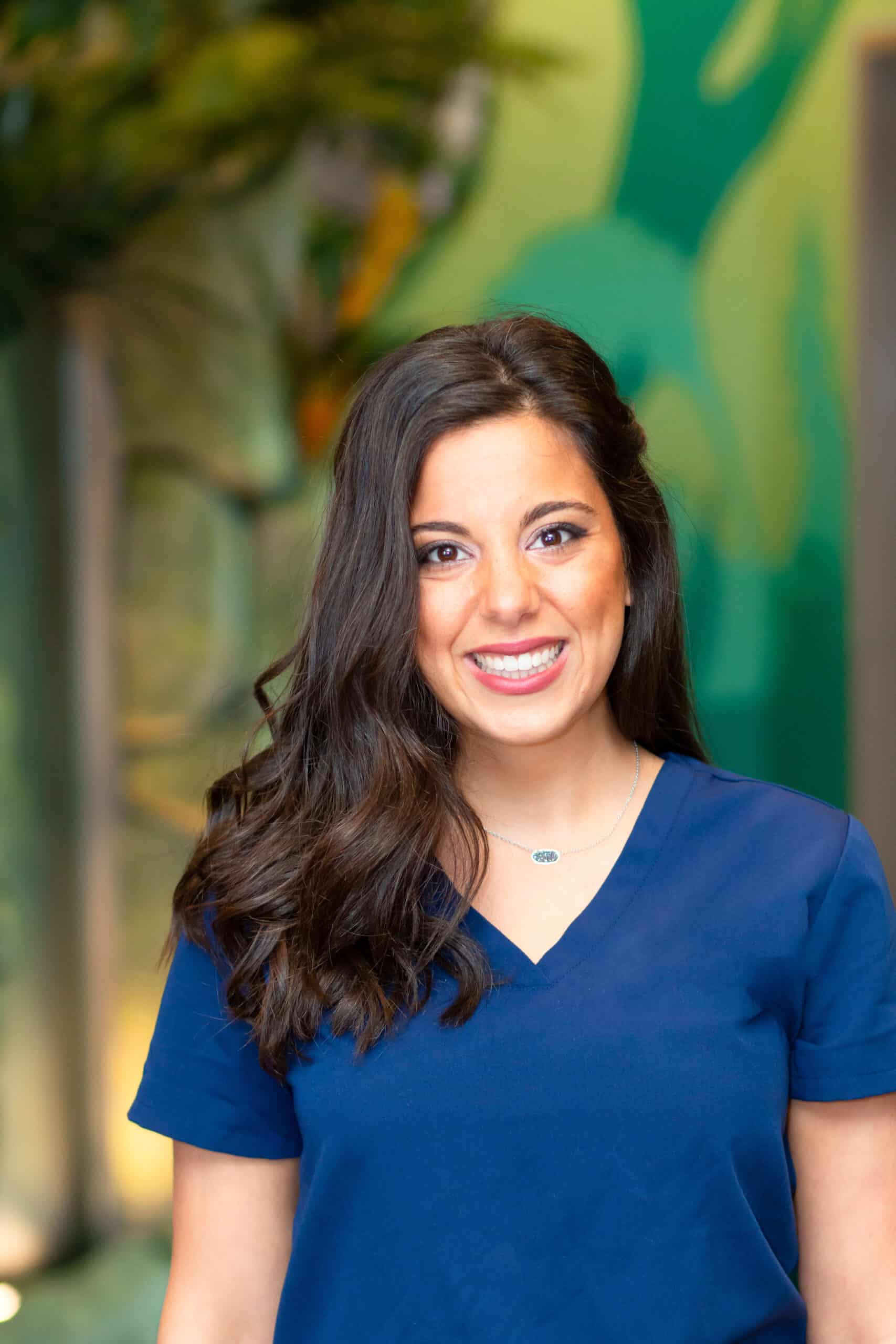 Smiling woman with long dark hair in blue scrubs, standing in front of a colorful mural.